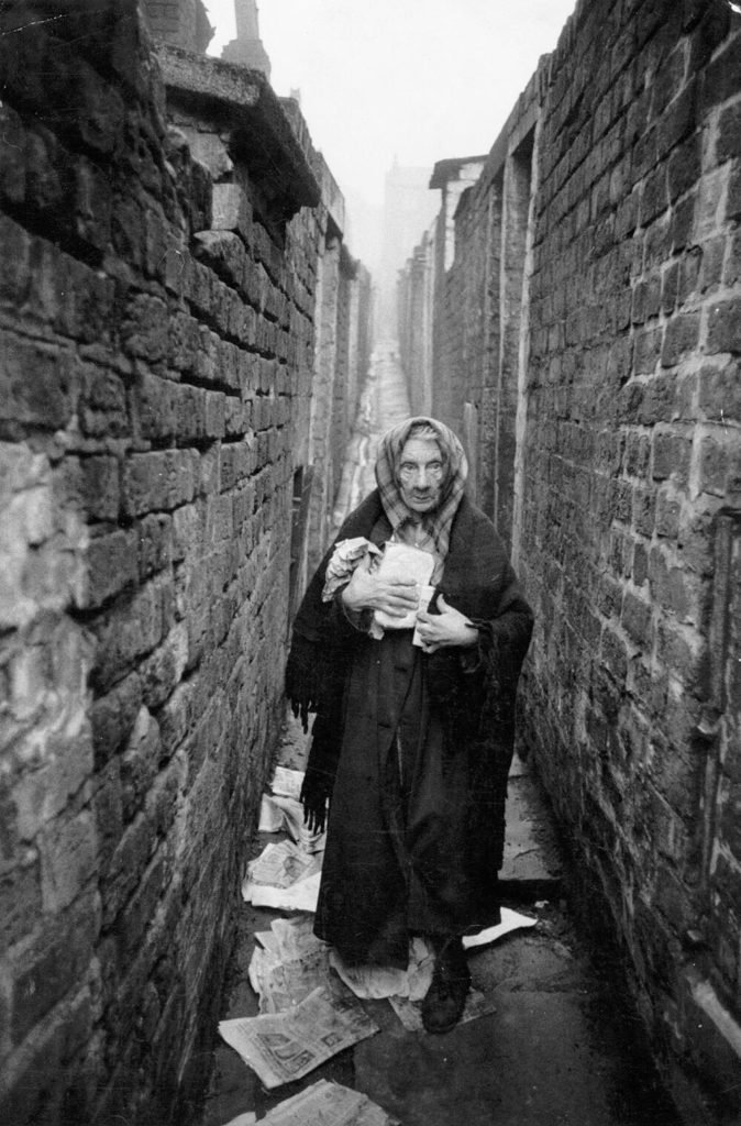 170.Thurston Hopkins - Lo sguardo umano della fotografia britannica 27 26.Una donna anziana in piedi tra i rifiuti in un vicolo dei bassifondi di Liverpool. Liverpool Slums reportage 1956. Thurston Hopkins
