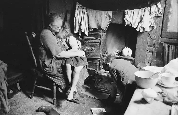170.Thurston Hopkins - Lo sguardo umano della fotografia britannica 30 29.Liverpool Slum reportage1956. Una donna seduta vicino a una stufa con due bambini nella loro casa nella baraccopoli di Frank Street a Liverpool.Thurston Hopkins